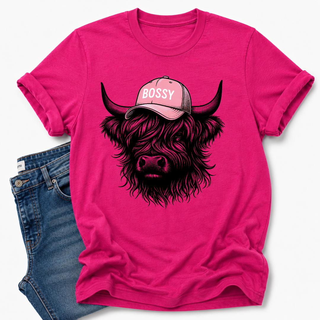 A bright pink short-sleeve t-shirt with a graphic of a shaggy Highland cow wearing a pink cap that says 'BOSSY' in white letters. The t-shirt is styled flat-lay with a pair of blue jeans on a clean white background.  Grab your tee from Coffee and Moo NZ.