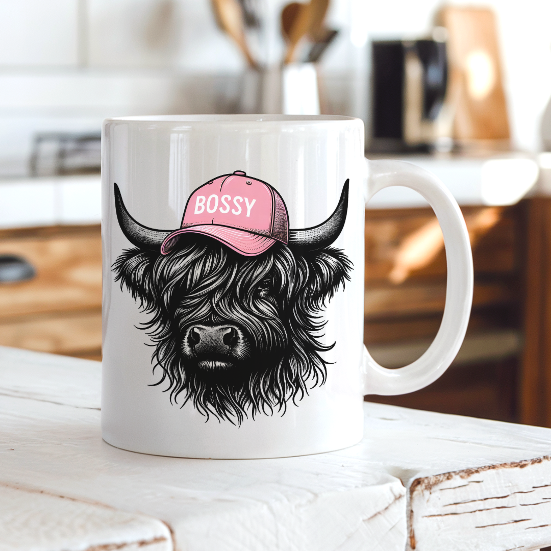 A white ceramic mug featuring a black illustration of a shaggy Highland cow wearing a pink cap with the word 'BOSSY' in white, set on a rustic white wooden table with a blurred kitchen background. grab your mug from Coffee and Moo NZ.