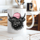 A white ceramic mug featuring a black illustration of a shaggy Highland cow wearing a pink cap with the word 'BOSSY' in white, set on a rustic white wooden table with a blurred kitchen background. grab your mug from Coffee and Moo NZ.