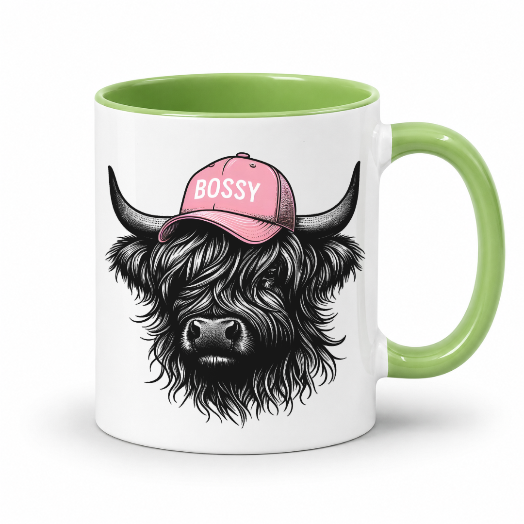 A white ceramic mug with a light green handle and interior, featuring a  of a black shaggy Highland cow wearing a pink cap with the word 'BOSSY' in white text. grab your bossy  mug from Coffee and Moo NZ.