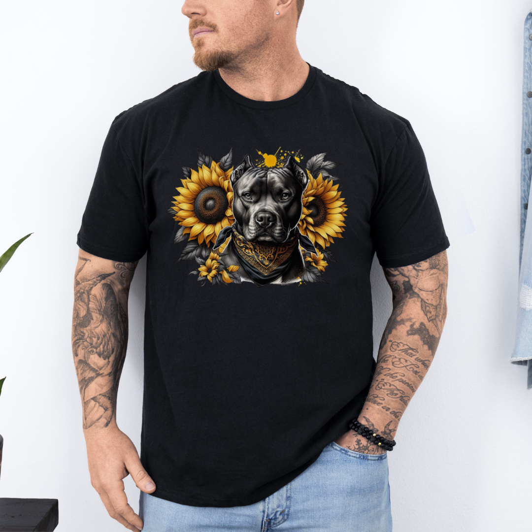 A man wearing a black short-sleeve graphic t-shirt featuring a detailed print of a pitbull dog wearing a bandana and surrounded by yellow sunflowers. The man has tattoos on both arms and is wearing light blue jeans, standing against a white wall. Grab your tee from coffee and moo NZ.