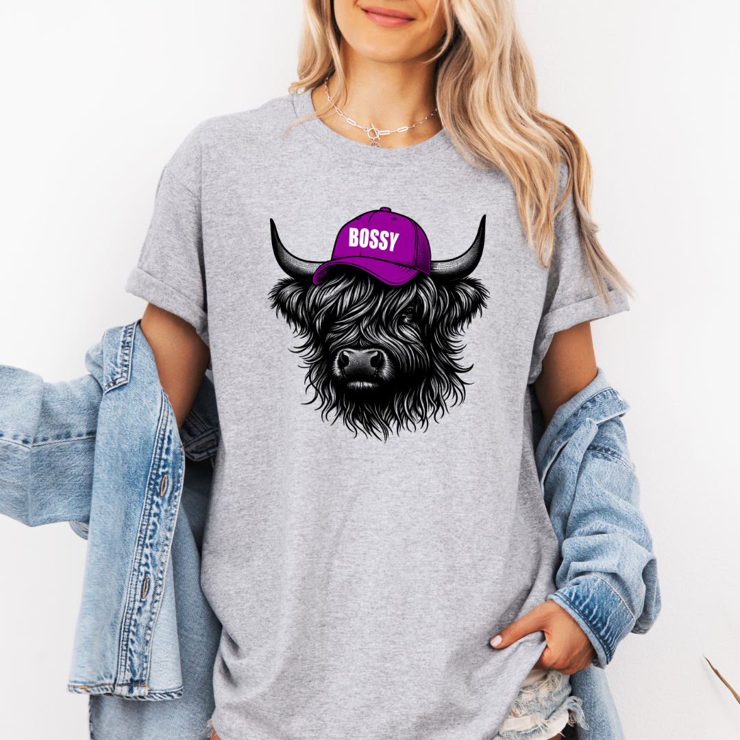 A woman wearing a heather gray oversized t-shirt with a graphic of a black Scottish Highland cow wearing a purple cap that says 'BOSSY' in white. She has a light blue denim jacket draped off her shoulders against a plain white background. Grab your Bossy highland coo tee from Coffee and Moo NZ.