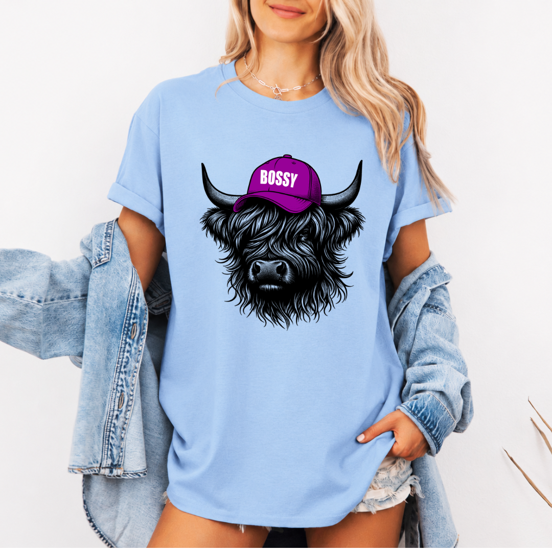 A woman wearing a light blue oversized graphic t-shirt with a black highland cow illustration wearing a purple cap that says 'BOSSY'. She has a light-wash denim jacket off her shoulders and is posing against a clean white background. Grab your Bossy highland coo tee from Coffee and Moo NZ.