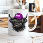 A white ceramic mug with a black and gray illustration of a shaggy Highland cow wearing a purple cap that says 'BOSSY' in white letters, set on a rustic white wooden table with a blurred kitchen background. Grab your bossy highland coo mug from Coffee and Moo NZ.