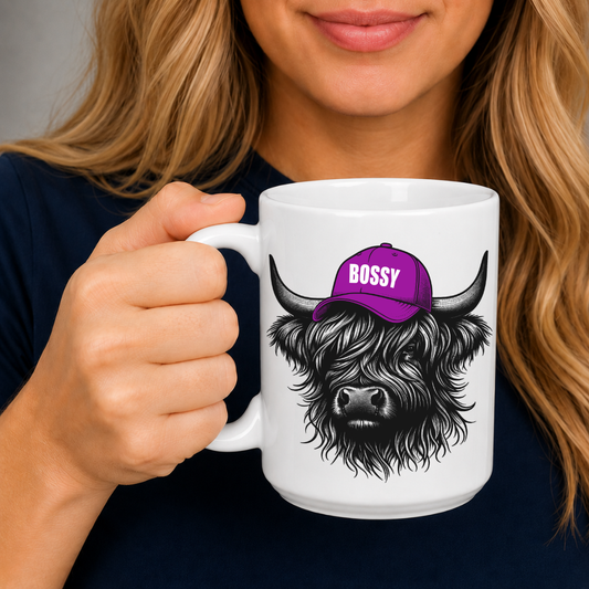 A person holding a white ceramic mug with a black-and-gray highland cow illustration wearing a purple cap that says 'BOSSY' in white letters. Grab your bossy highland coo mug from Coffee and Moo NZ.