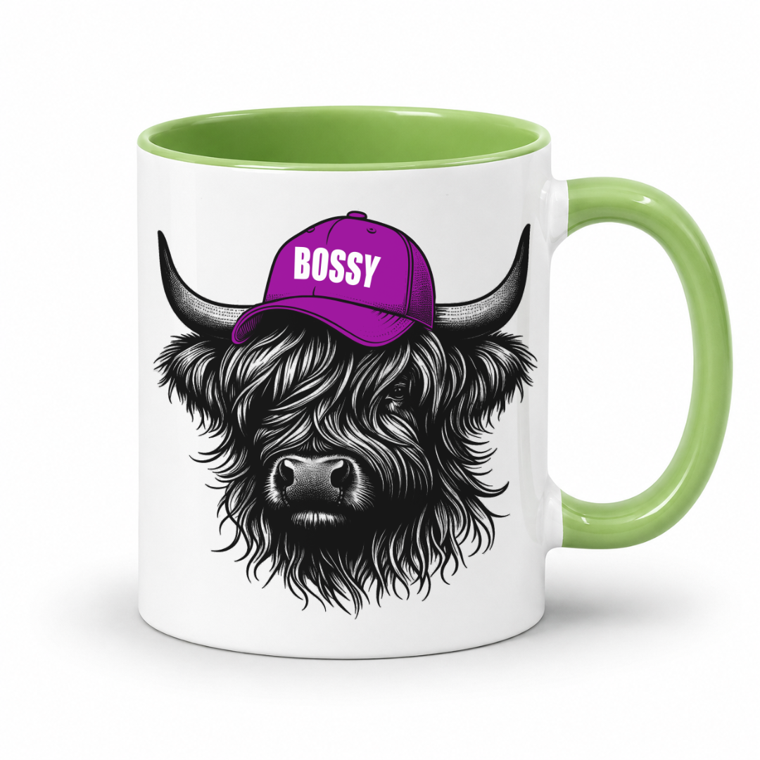 A white ceramic mug with a light green interior and handle, featuring a graphic of a shaggy Highland cow wearing a purple cap with the word 'BOSSY' in white. Grab your bossy highland coo mug from Coffee and Moo NZ.