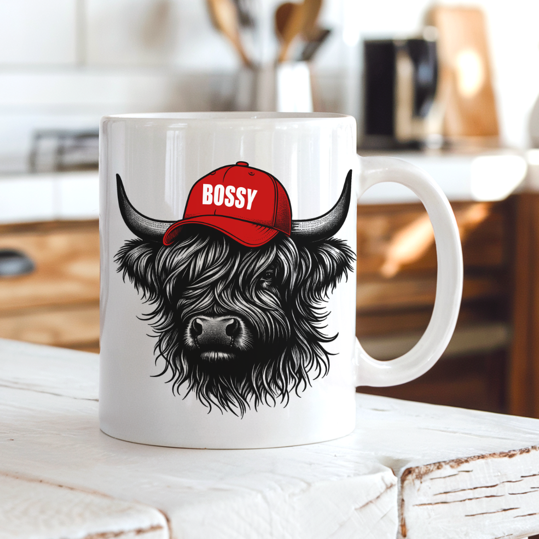 A white ceramic mug featuring a black and white illustration of a shaggy Highland cow wearing a red baseball cap with the word 'BOSSY' in white, set on a rustic white wooden table with a blurred kitchen background. Grab your Bossy highland coo mug from Coffee and Moo NZ.