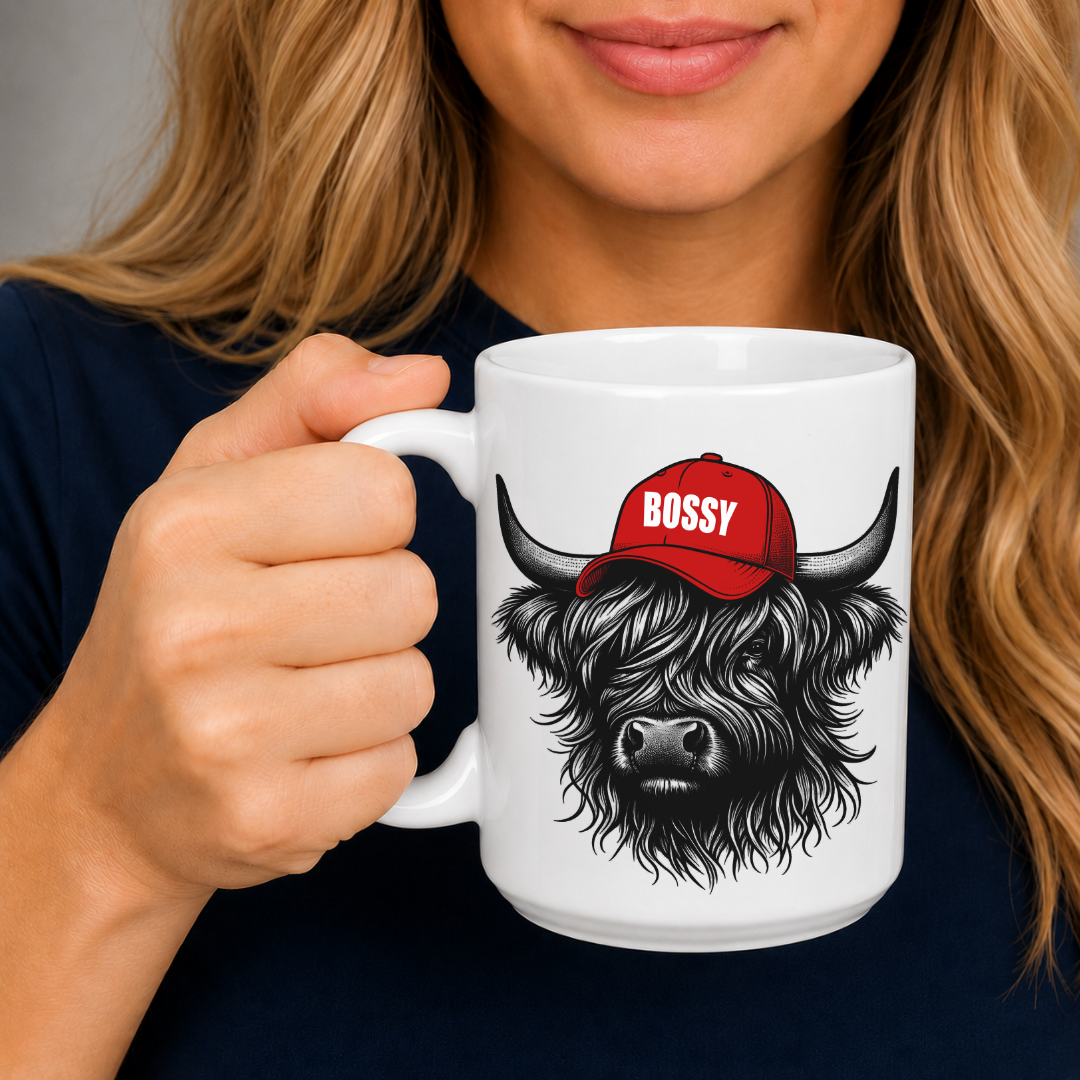 A close-up of a woman holding a white ceramic mug with a black-and-white highland cow illustration wearing a red cap that says "BOSSY.". Grab your Bossy highland coo mug from Coffee and Moo NZ.