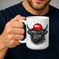 A person holding a white ceramic mug with a graphic of a shaggy black-and-white Highland cow wearing a red cap with the word 'BOSSY' in white, against a dark navy shirt and grey background. Grab your Bossy highland coo mug from Coffee and Moo NZ.