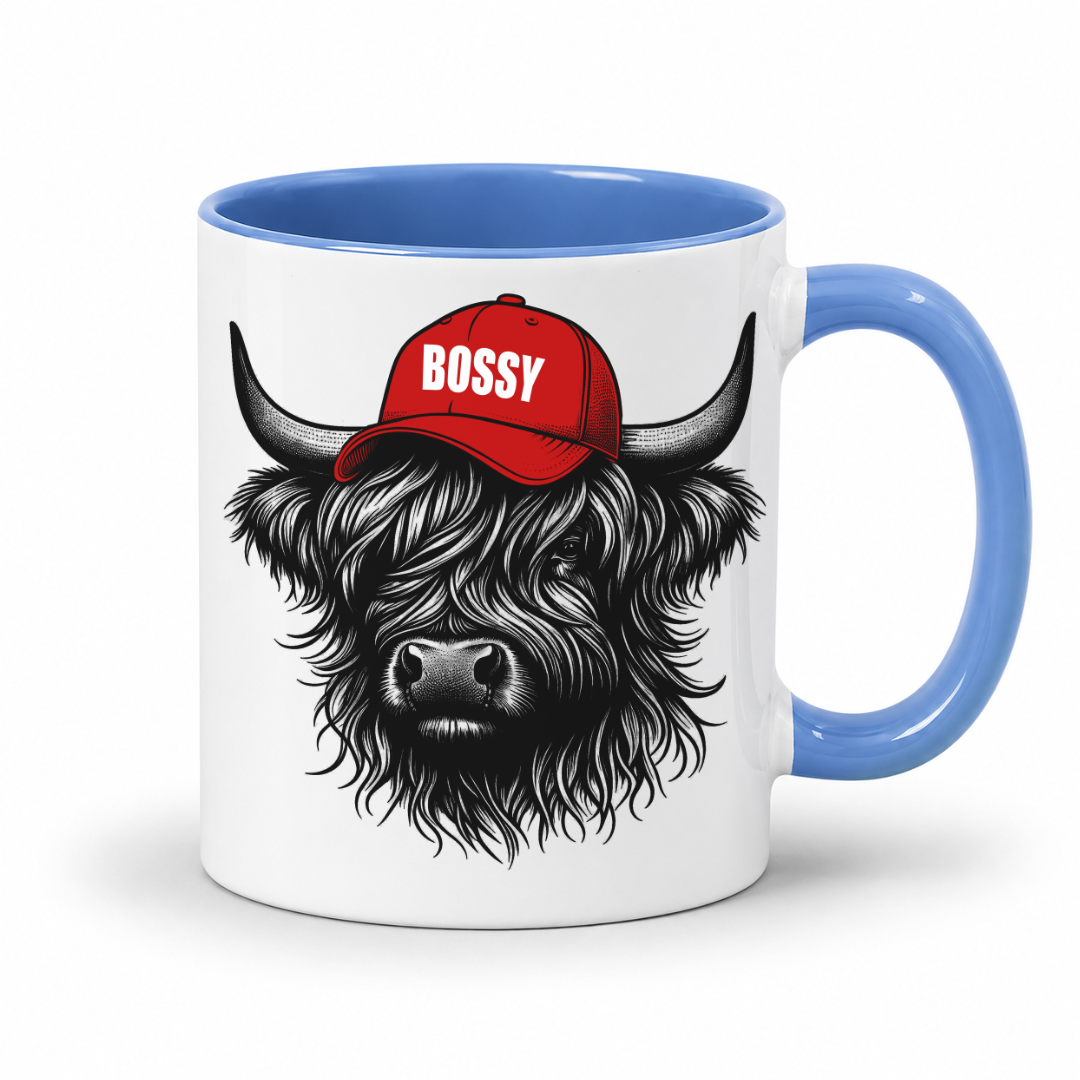 A white ceramic mug with a blue handle and interior, featuring a graphic of a shaggy black highland cow wearing a red cap with the word 'BOSSY' in white. Grab your Bossy highland coo mug from Coffee and Moo NZ.