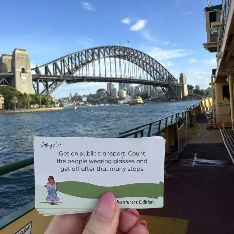 The Getting Lost Solo Adventurers Edition is your ultimate permit to escape the noise, skip the group compromises, and explore the world entirely at your own pace.  The card reads, 'Getting Lost' with a small illustration of a girl walking on green hills. Grab your game from Coffee and Moo NZ