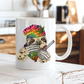 A white ceramic mug featuring a colorful illustration of a sloth with red eyes, wearing a rainbow tie-dye beanie and holding a bong and baggy. set on a rustic white wooden table with a blurred kitchen background. Grab your mug from Coffee & Moo NZ, 