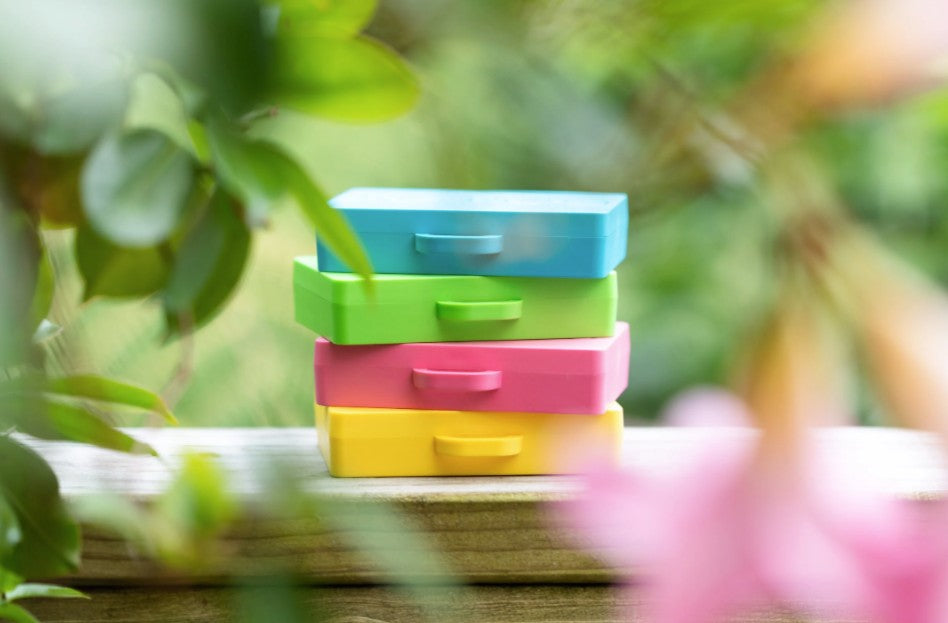 A stack of four small rectangular suitcases for the "Getting Lost" game in blue, green, pink, and yellow, placed on a wooden surface with blurred green leaves and pink flowers in the foreground and background. Purchase from Coffee and Moo NZ.