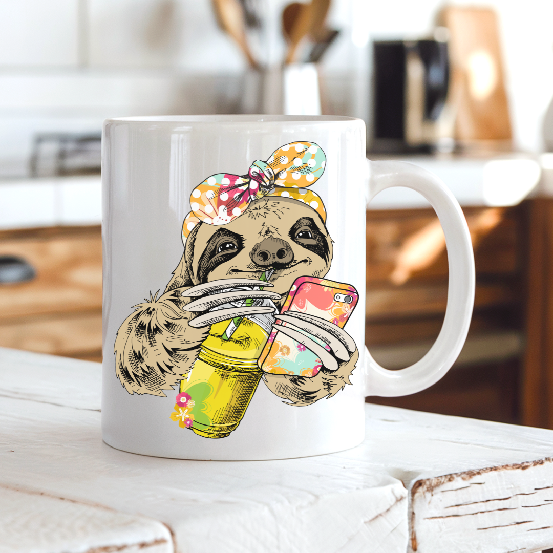A white ceramic mug with a colorful illustration of a sloth wearing a polka-dot bow bandana and holding a loaded-tea drink and a phone, set on a rustic white wooden table with a blurred kitchen background. Grab yours from Coffee and Moo NZ.
