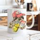 A white ceramic mug with a colorful illustration of a sloth wearing a coral coloured cap and holding a loaded-tea drink and a phone, set on a rustic white wooden table with a blurred kitchen background. Grab yours from Coffee and Moo NZ.