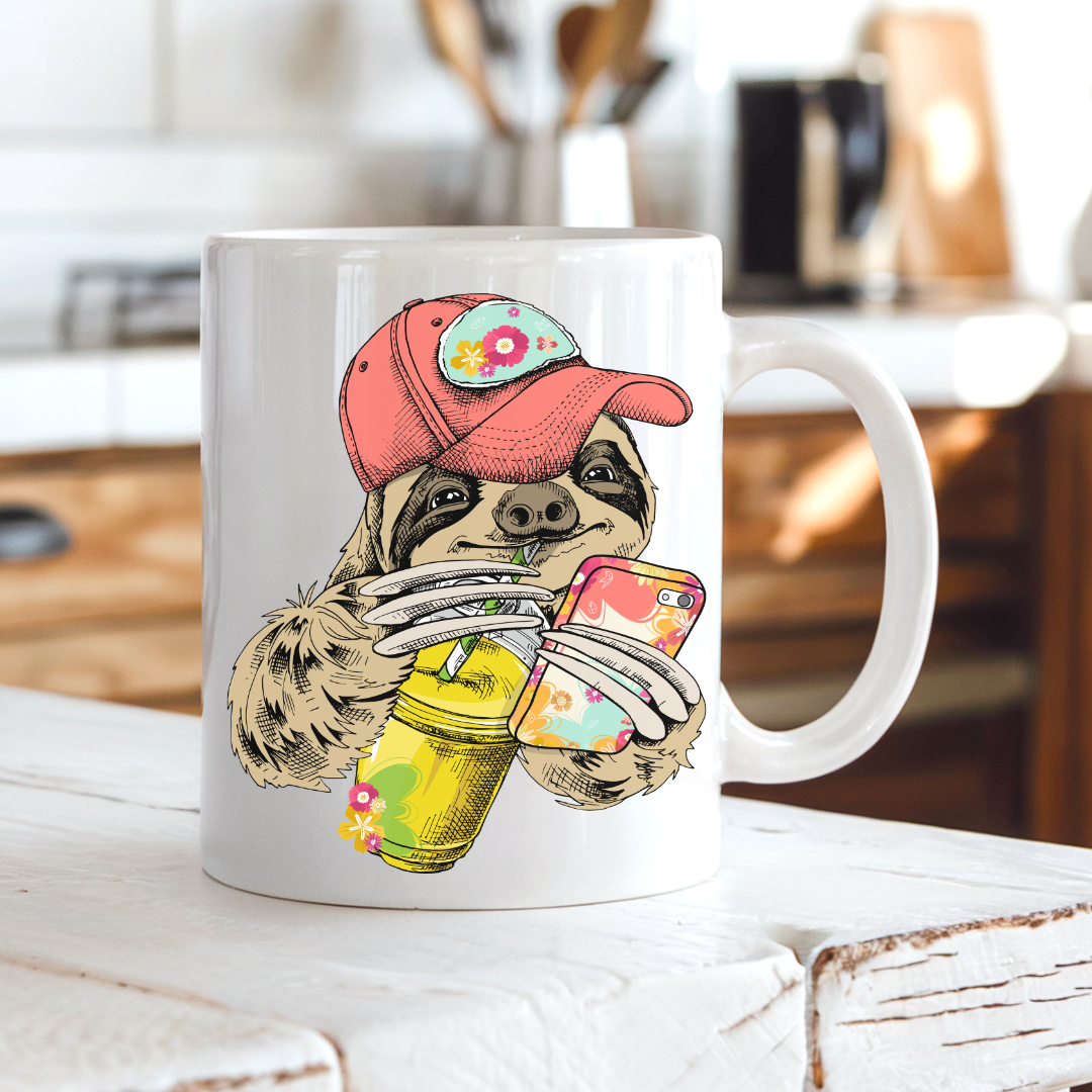 A white ceramic mug with a colorful illustration of a sloth wearing a coral coloured cap and holding a loaded-tea drink and a phone, set on a rustic white wooden table with a blurred kitchen background. Grab yours from Coffee and Moo NZ.