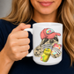 A person holding a white ceramic mug  mug with a colorful illustration of a sloth wearing a coral coloured cap and holding a loaded-tea drink and a phone. Grab yours from Coffee and Moo NZ.
