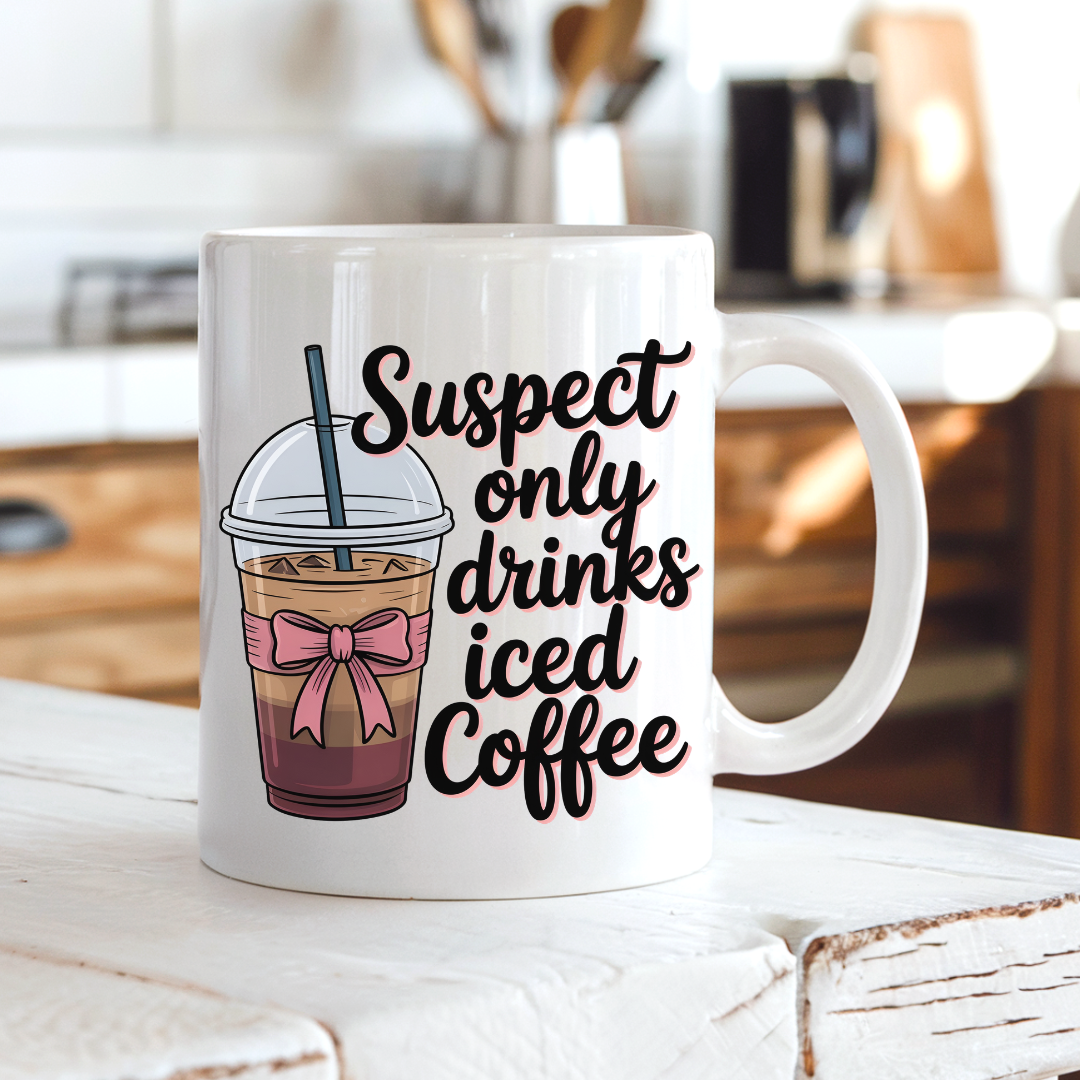 A white ceramic coffee mug with a handle, featuring a graphic of an iced coffee cup with a pink bow and the text 'Suspect only drinks iced Coffee' in black script, set on a rustic white wooden table with a blurred kitchen background. From Coffee and Moo NZ.