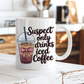 A white ceramic coffee mug with a handle, featuring a graphic of an iced coffee cup with a pink bow and the text 'Suspect only drinks iced Coffee' in black script, set on a rustic white wooden table with a blurred kitchen background. From Coffee and Moo NZ.