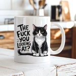 A white ceramic coffee mug with a black and white tuxedo cat illustration and the text 'THE FUCK YOU LOOKING AT?' printed on the side, set on a rustic white wooden table with a blurred kitchen background. From Coffee and Moo