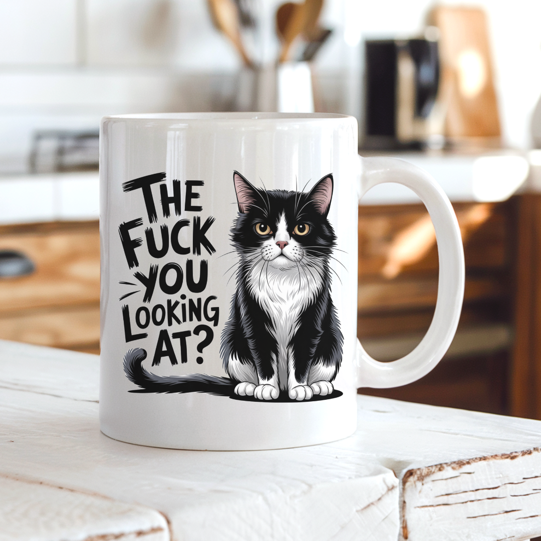 A white ceramic coffee mug with a black and white tuxedo cat illustration and the text 'THE FUCK YOU LOOKING AT?' printed on the side, set on a rustic white wooden table with a blurred kitchen background. From Coffee and Moo