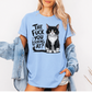 A woman wearing a light blue graphic t-shirt with a black and white cat illustration and the text 'THE FUCK YOU LOOKING AT?'. She has a light-wash denim jacket off her shoulders and is posing against a clean white background. Grab your feral tee from Coffee and Moo NZ.