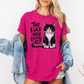 A woman wearing a bright pink graphic t-shirt with a black and white cat illustration and the text 'THE FUCK YOU LOOKING AT?'. She has a light blue denim jacket draped off her shoulders against a plain white background. Grab your feral tee from Coffee and Moo NZ.