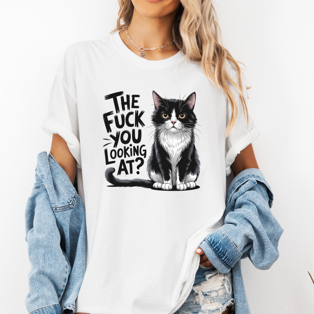 A woman wearing a white graphic t-shirt with a black and white cat illustration and the text 'THE FUCK YOU LOOKING AT?'. She has a light blue denim shirt off her shoulders and is wearing distressed jeans against a plain white background. Grab your feral tee from Coffee and Moo NZ.