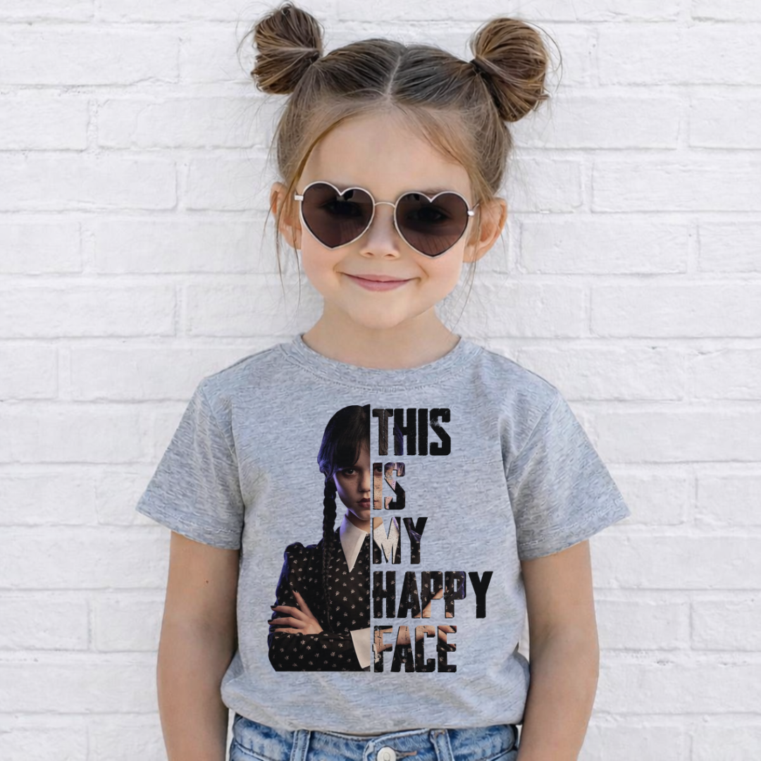 A young girl with her hair in two buns and heart-shaped sunglasses is wearing a heather gray graphic t-shirt featuring a collage of Wednesday Addams and the text 'THIS IS MY HAPPY FACE'. She is standing against a white painted brick wall. Grab your Kids tee from Coffee and Moo NZ