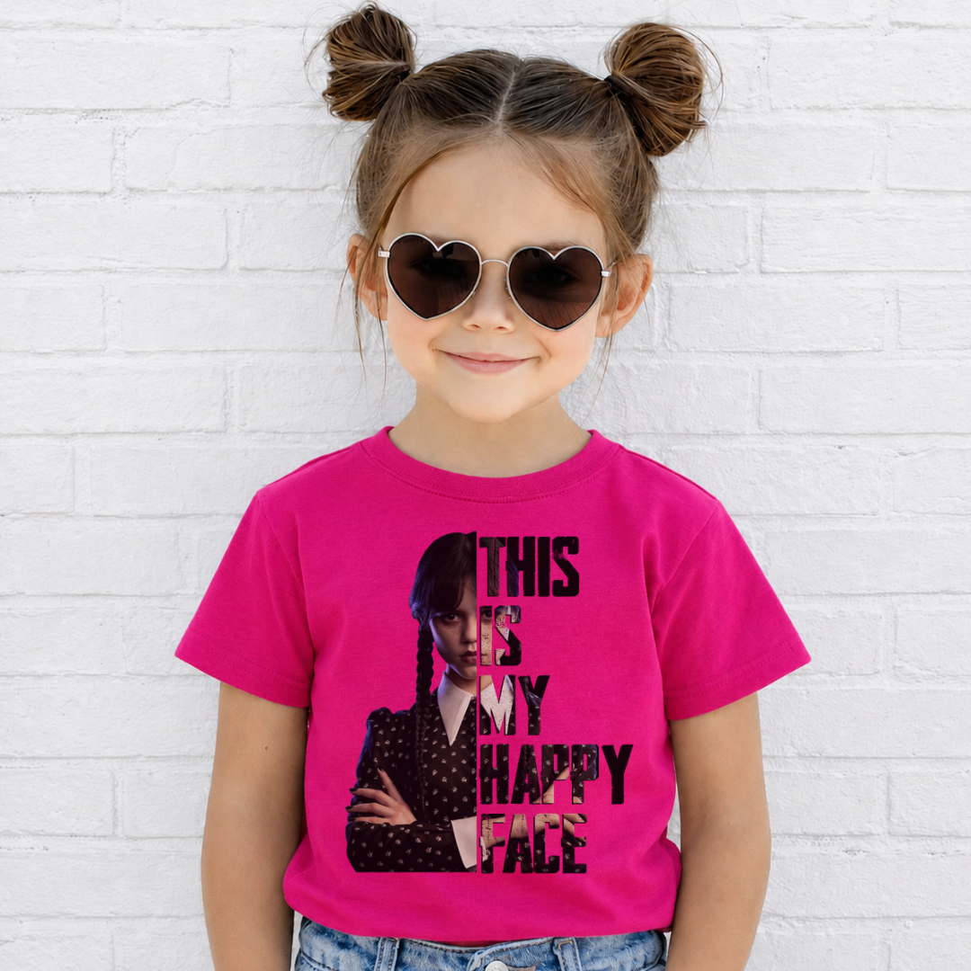 A young girl with brown hair in two buns and heart-shaped sunglasses wears a bright pink graphic t-shirt featuring Wednesday Addams and the text 'THIS IS MY HAPPY FACE'. She is standing against a white painted brick wall. Grab your Kids tee from Coffee and Moo NZ