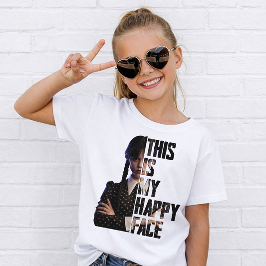 A young girl wearing a white graphic t-shirt with a black and brown print of Wednesday Addams and the text 'THIS IS MY HAPPY FACE', posing with heart-shaped sunglasses against a white brick wall. Grab your Kids tee from Coffee and Moo NZ