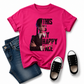 A bright pink graphic T-shirt laid flat on a white background, styled with folded blue jeans and a pair of black low-top sneakers. The shirt features a printed portrait of Wednesday Addams and the text 'THIS IS MY HAPPY FACE' in black. Grab your Kids tee from Coffee and Moo NZ