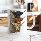 A white ceramic mug with a printed graphic of three Highland cattle heads stacked vertically against a fence post, set on a rustic white wooden table in a blurred kitchen background.  From Coffee and Moo NZ