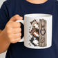 A child holding a white ceramic mug with a printed illustration of three horses peeking from behind a wooden fence, wearing a navy blue shirt. From Coffee and Moo