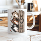 A white ceramic coffee mug with a printed illustration of three horses peeking from behind a rustic wooden fence with a heart-shaped ornament, set on a white wooden table in a blurred kitchen background. From Coffee and Moo