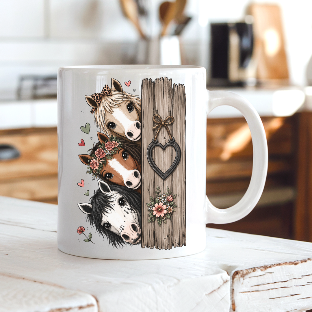 A white ceramic coffee mug with a printed illustration of three horses peeking from behind a rustic wooden fence with a heart-shaped ornament, set on a white wooden table in a blurred kitchen background. From Coffee and Moo