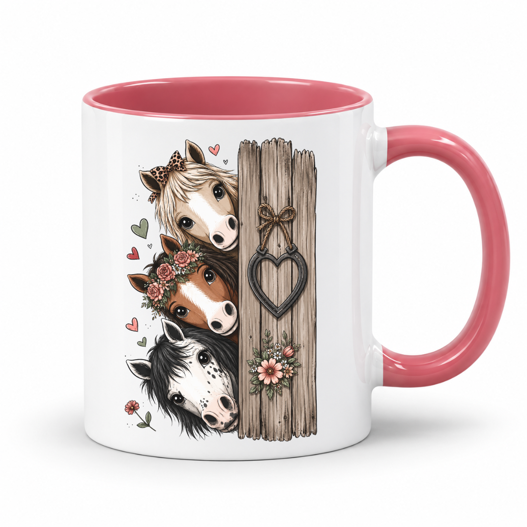 A white ceramic mug with a pink interior and handle, featuring a printed graphic of three cartoon horses beside a rustic wooden fence with a heart-shaped ornament. From Coffee and Moo