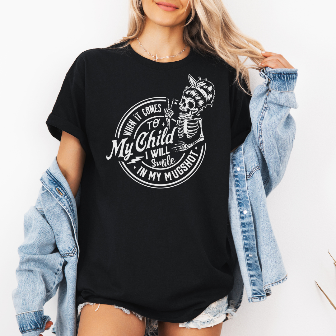 A woman wearing a black graphic t-shirt with a white skeleton and text design Saying "when it comes to my child, i will smile in my mugshot" styled with an open light blue denim jacket and denim shorts. Purchase from Coffee and Moo NZ.