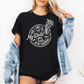 A woman wearing a black graphic t-shirt with a white skeleton and text design Saying "when it comes to my child, i will smile in my mugshot" styled with an open light blue denim jacket and denim shorts. Purchase from Coffee and Moo NZ.