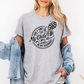 A woman wearing a grey graphic t-shirt with a white skeleton and text design Saying "when it comes to my child, i will smile in my mugshot" styled with an open light blue denim jacket and denim shorts. Purchase from Coffee and Moo NZ.