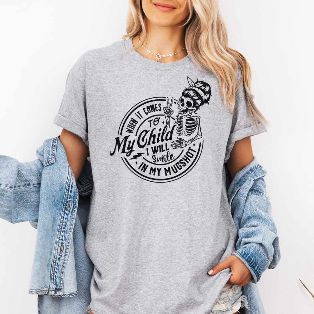 A woman wearing a grey graphic t-shirt with a white skeleton and text design Saying "when it comes to my child, i will smile in my mugshot" styled with an open light blue denim jacket and denim shorts. Purchase from Coffee and Moo NZ.