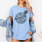 A woman wearing a light blue graphic t-shirt with a white skeleton and text design Saying "when it comes to my child, i will smile in my mugshot" styled with an open light blue denim jacket and denim shorts. Purchase from Coffee and Moo NZ.