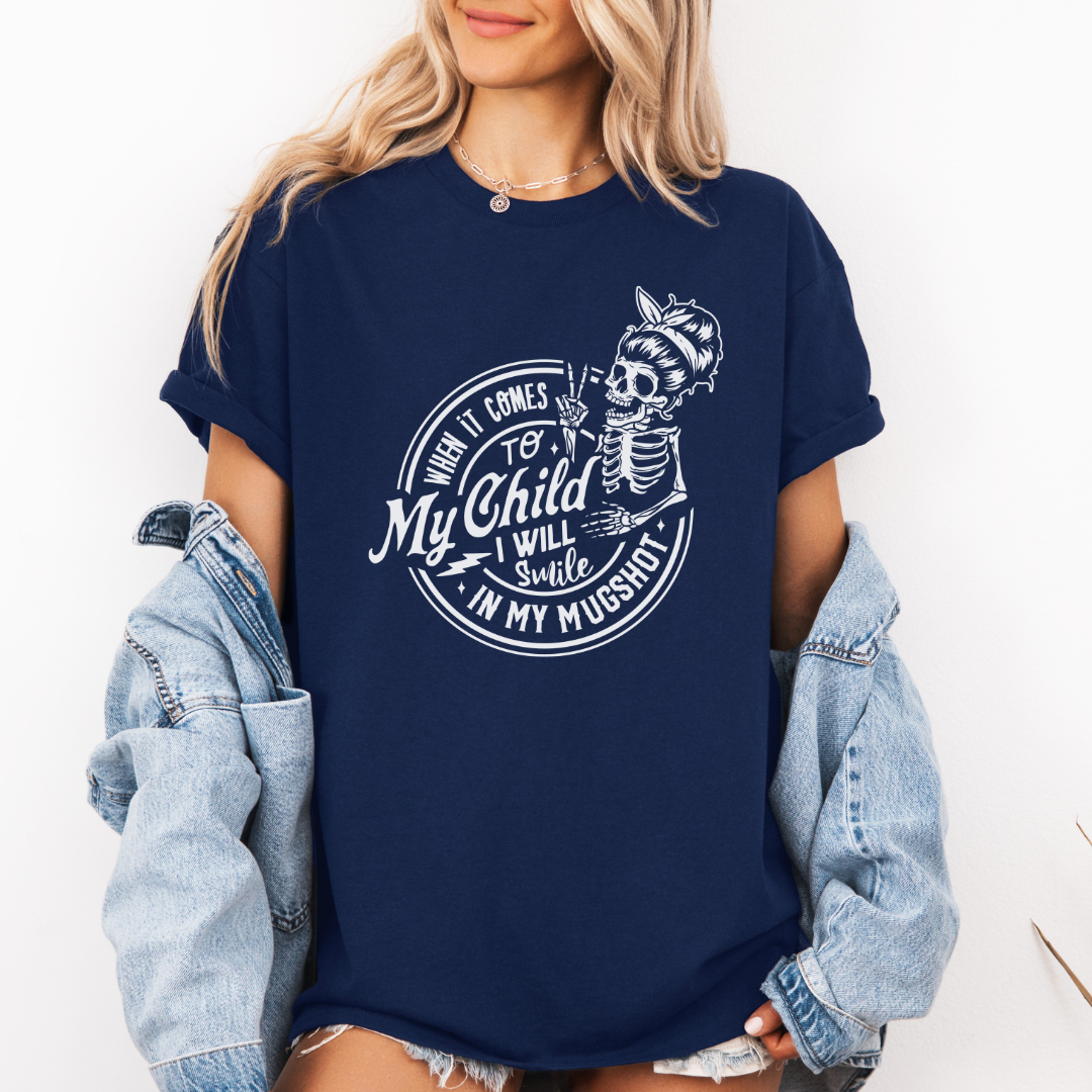 A woman wearing a navy graphic t-shirt with a white skeleton and text design Saying "when it comes to my child, i will smile in my mugshot" styled with an open light blue denim jacket and denim shorts. Purchase from Coffee and Moo NZ.