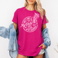 A woman wearing a bright pink graphic t-shirt with a white skeleton and text design Saying "when it comes to my child, i will smile in my mugshot" styled with an open light blue denim jacket and denim shorts. Purchase from Coffee and Moo NZ.