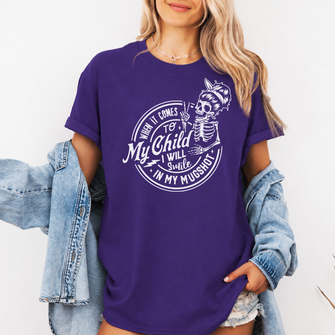 A woman wearing a purple graphic t-shirt with a white skeleton and text design Saying "when it comes to my child, i will smile in my mugshot" styled with an open light blue denim jacket and denim shorts. Purchase from Coffee and Moo NZ.