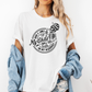 A woman wearing a white graphic t-shirt with a white skeleton and text design Saying "when it comes to my child, i will smile in my mugshot" styled with an open light blue denim jacket and denim shorts. Purchase from Coffee and Moo NZ.