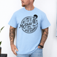 A man wearing a light blue graphic t-shirt with a white skeleton and text design Saying "when it comes to my child, i will smile in my mugshot" styled with blue denim jeans. Purchase from Coffee and Moo NZ.