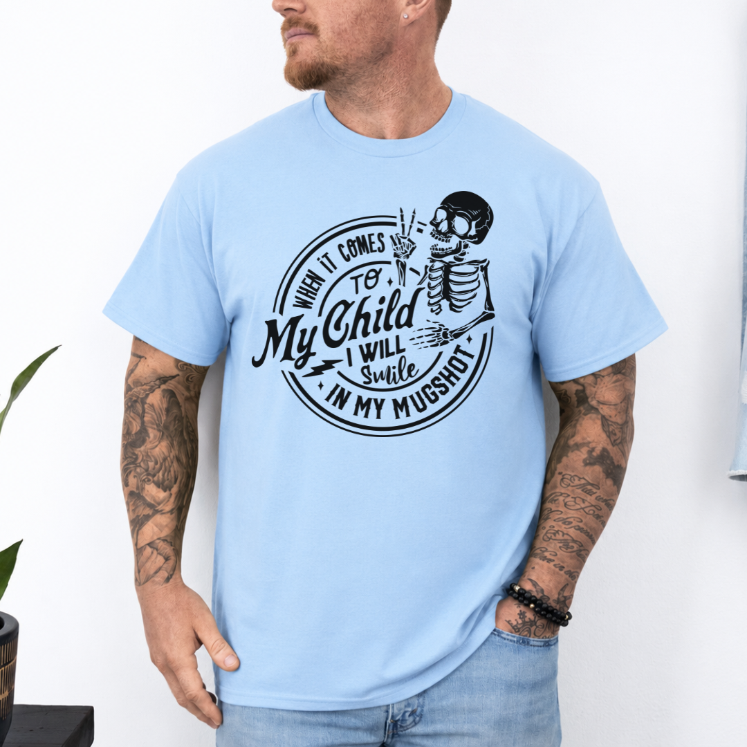 A man wearing a light blue graphic t-shirt with a white skeleton and text design Saying "when it comes to my child, i will smile in my mugshot" styled with blue denim jeans. Purchase from Coffee and Moo NZ.