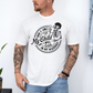 A man wearing a white graphic t-shirt with a white skeleton and text design Saying "when it comes to my child, i will smile in my mugshot" styled with blue denim jeans. Purchase from Coffee and Moo NZ.