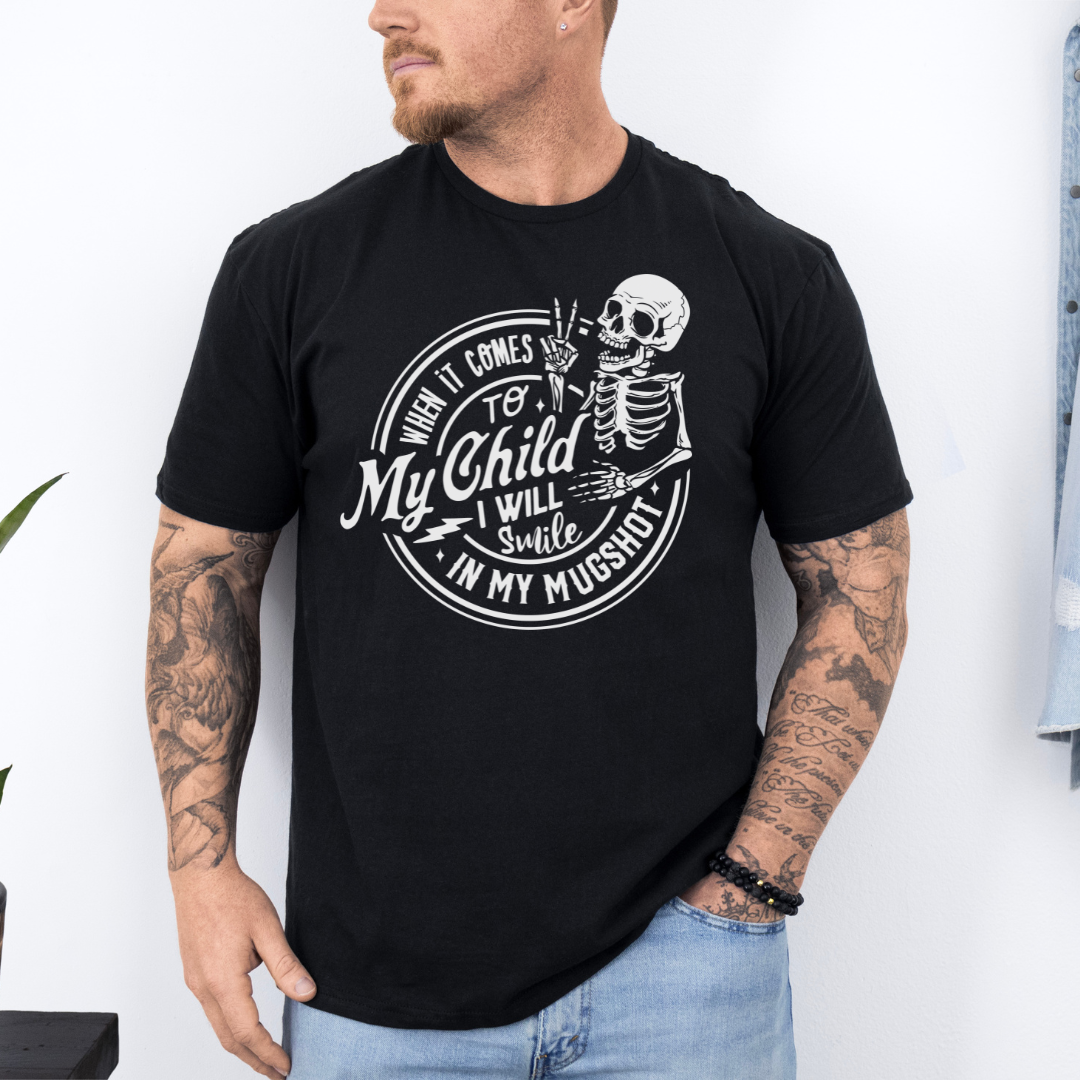 A man wearing a black graphic t-shirt with a white skeleton and text design Saying "when it comes to my child, i will smile in my mugshot" styled with blue denim jeans. Purchase from Coffee and Moo NZ.